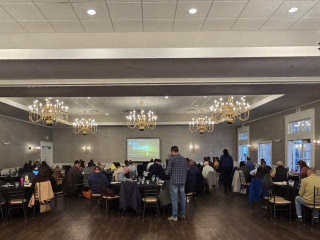 Attendees seated at round tables facing a presentation screen at the AMWS annual meeting.